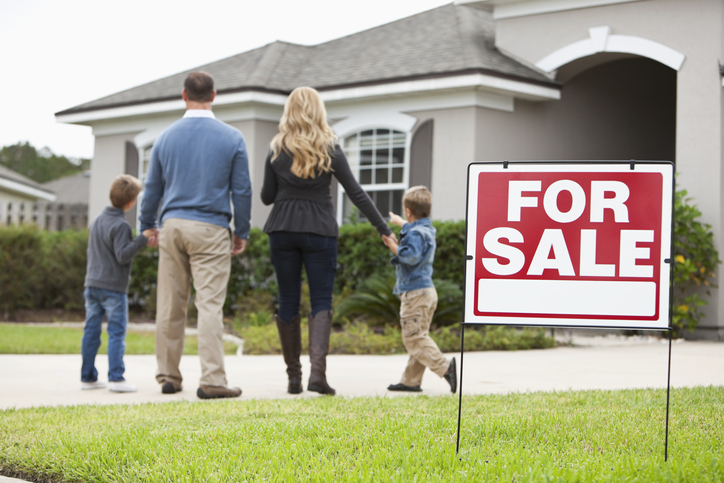 Family with two boys (4 and 6 years) standing in front of house with FOR SALE sign in front yard. Focus on sign.