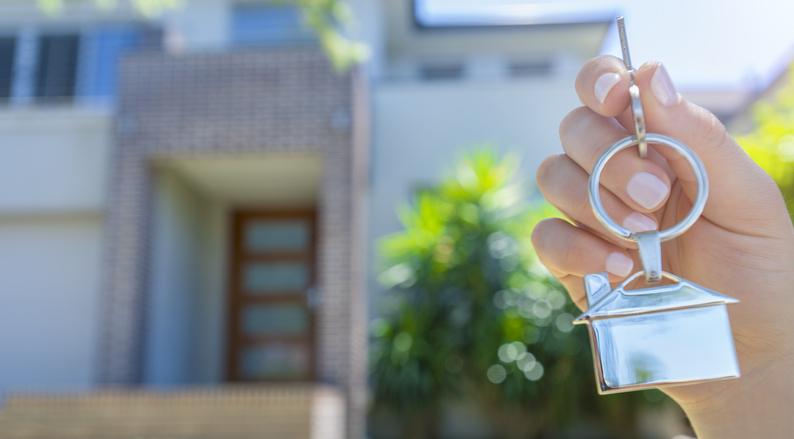 Hand holding a house key in front of a large house. The key ring has a house shaped icon on the end and is shiny silver colour. The house has a tree in front of it and you can see the front door in the background. The key ring house icon is dangling down. Close up with shallow focus and copy space. New house concept.